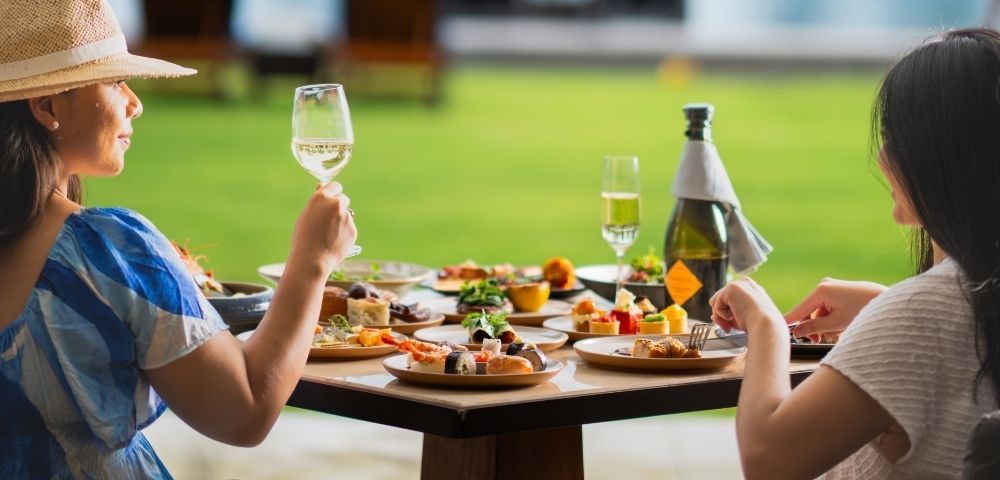 Two women enjoying a vibrant outdoor meal at a table laden with assorted dishes. One raises a glass of white wine, conveying a relaxed, joyful atmosphere.