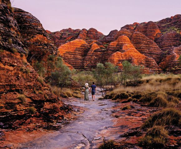 Two people walk on a rocky path surrounded by striking orange, layered rock formations. The scene conveys adventure and natural beauty.