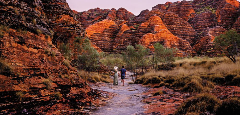 Two people walk on a rocky path surrounded by striking orange, layered rock formations. The scene conveys adventure and natural beauty.