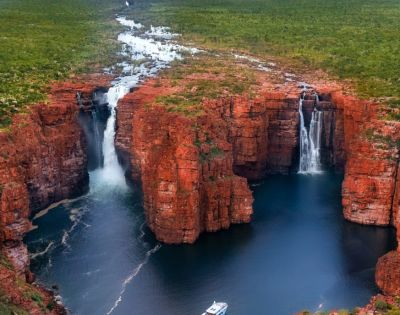 Aerial view of twin waterfalls cascading over rugged, red rock cliffs into a deep blue pool. Lush greenery surrounds the scene, creating a serene atmosphere.