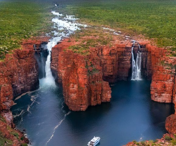 Aerial view of twin waterfalls cascading over rugged, red rock cliffs into a deep blue pool. Lush greenery surrounds the scene, creating a serene atmosphere.