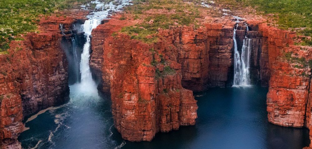 Aerial view of twin waterfalls cascading over rugged, red rock cliffs into a deep blue pool. Lush greenery surrounds the scene, creating a serene atmosphere.