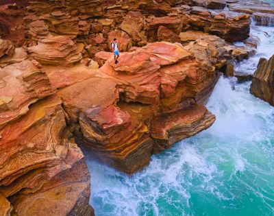 A person stands on striking red and orange layered cliffs, overlooking vibrant turquoise waves crashing against the rocks, evoking adventure and serenity.