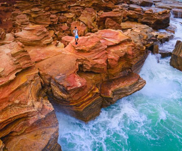 A person stands on striking red and orange layered cliffs, overlooking vibrant turquoise waves crashing against the rocks, evoking adventure and serenity.