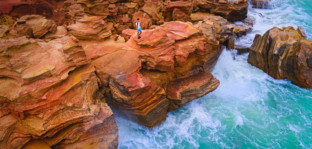 A person stands on striking red and orange layered cliffs, overlooking vibrant turquoise waves crashing against the rocks, evoking adventure and serenity.