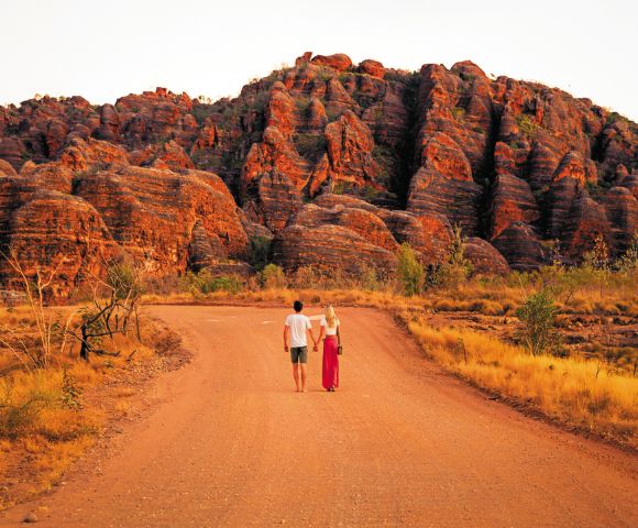 A couple walks hand in hand down a reddish dirt road toward a unique sandstone rock formation under a clear sky, evoking adventure and serenity.