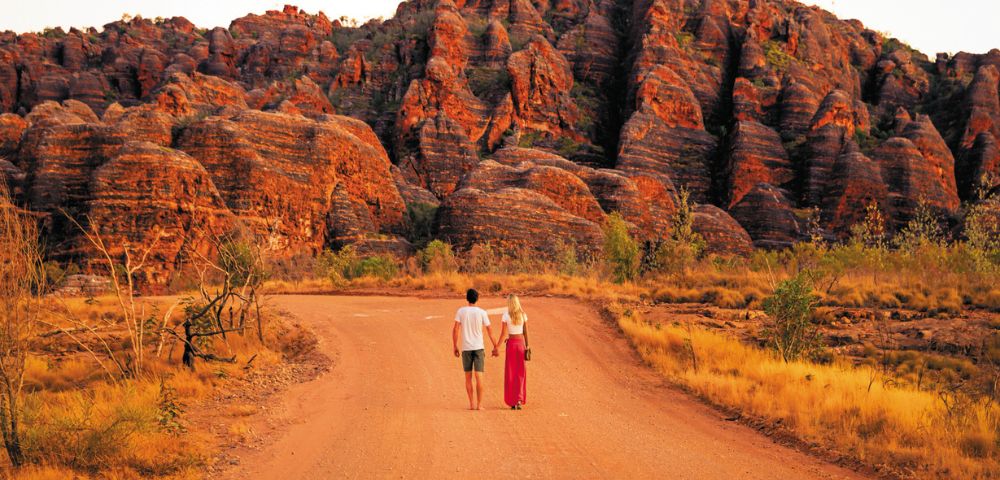 A couple walks hand in hand down a reddish dirt road toward a unique sandstone rock formation under a clear sky, evoking adventure and serenity.