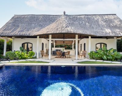A bright blue swimming pool in the foreground reflects the sky, with a thatched-roof building featuring an open-air lounge area in the background.
