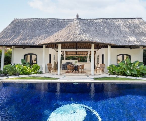 A bright blue swimming pool in the foreground reflects the sky, with a thatched-roof building featuring an open-air lounge area in the background.