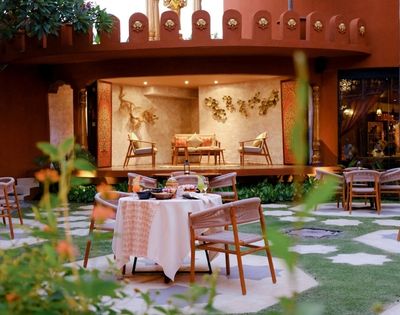An outdoor courtyard dining area with a table set for a meal, surrounded by chairs, in front of a raised platform with seating and ornate wall decorations