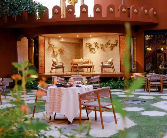 An outdoor courtyard dining area with a table set for a meal, surrounded by chairs, in front of a raised platform with seating and ornate wall decorations