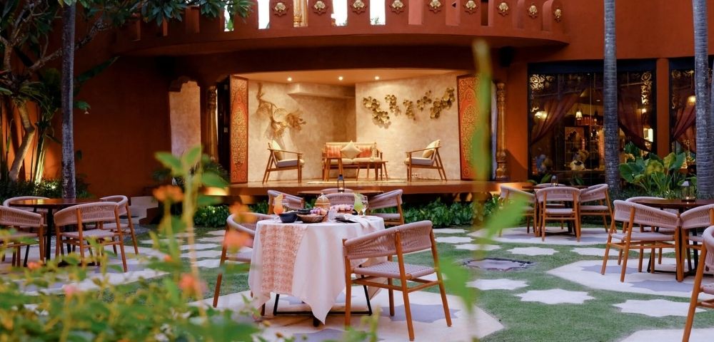 An outdoor courtyard dining area with a table set for a meal, surrounded by chairs, in front of a raised platform with seating and ornate wall decorations