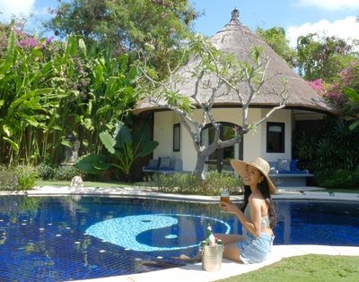 A woman in a sun hat sits by a blue swimming pool with a yin and yang symbol on the bottom, holding a drink in front of a thatched-roof bungalow surrounded by lush tropical plants