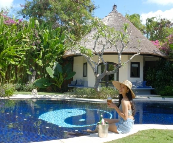A woman in a sun hat sits by a blue swimming pool with a yin and yang symbol on the bottom, holding a drink in front of a thatched-roof bungalow surrounded by lush tropical plants