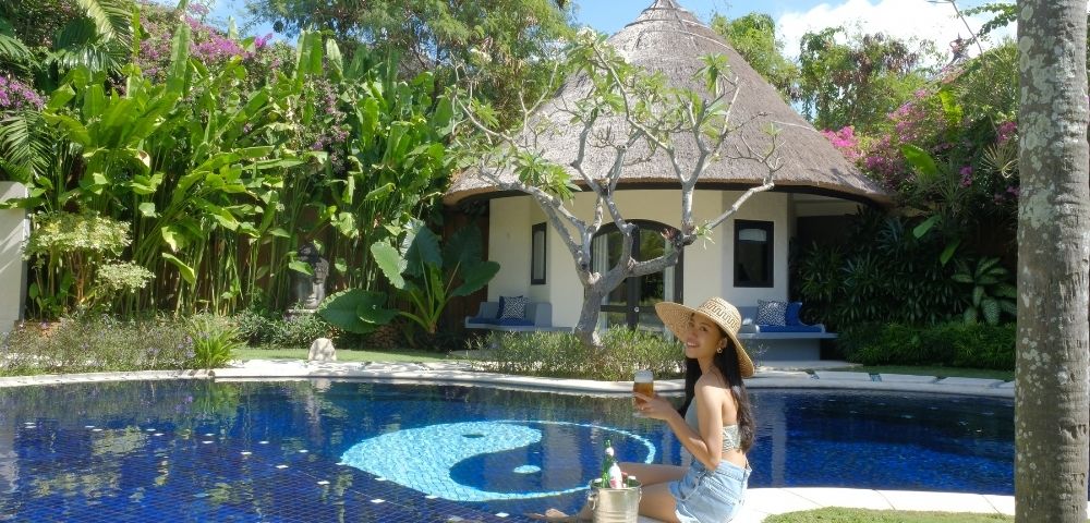 A woman in a sun hat sits by a blue swimming pool with a yin and yang symbol on the bottom, holding a drink in front of a thatched-roof bungalow surrounded by lush tropical plants
