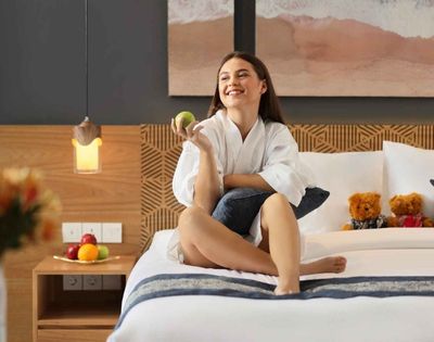 Smiling woman in a white robe sitting on a bed holding a green apple, with teddy bears and fruit on a nightstand in a cozy bedroom.