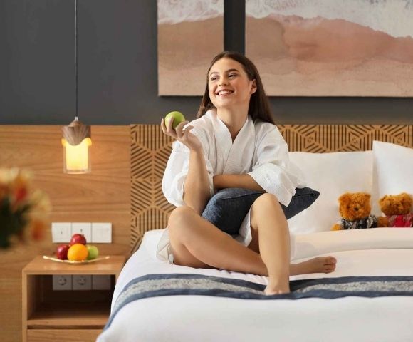 Smiling woman in a white robe sitting on a bed holding a green apple, with teddy bears and fruit on a nightstand in a cozy bedroom.