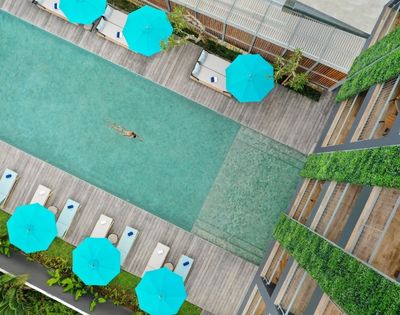Aerial view of a person swimming in a rectangular pool surrounded by lounge chairs, bright blue umbrellas, and a building with green walls.