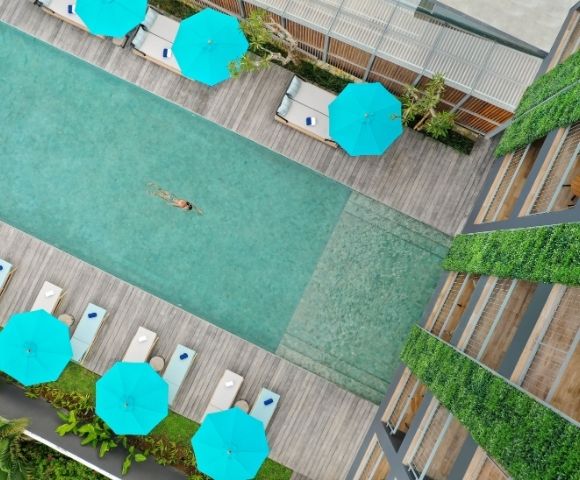 Aerial view of a person swimming in a rectangular pool surrounded by lounge chairs, bright blue umbrellas, and a building with green walls.