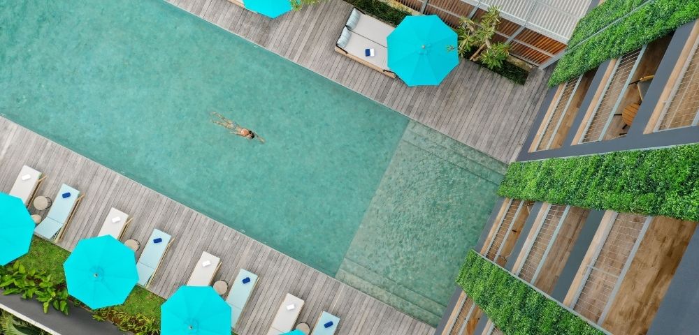 Aerial view of a person swimming in a rectangular pool surrounded by lounge chairs, bright blue umbrellas, and a building with green walls.