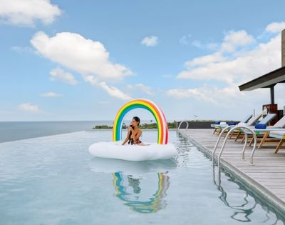 A person sits on a cloud-shaped inflatable with a rainbow arch in an infinity pool overlooking the ocean, with lounge chairs on the deck to the right under a partly cloudy sky.