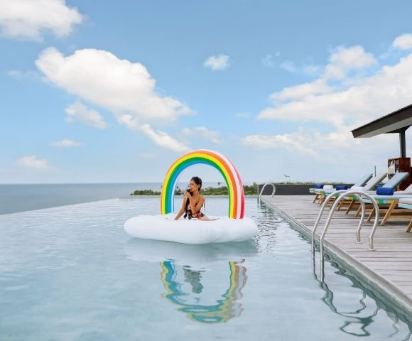 A person sits on a cloud-shaped inflatable with a rainbow arch in an infinity pool overlooking the ocean, with lounge chairs on the deck to the right under a partly cloudy sky.