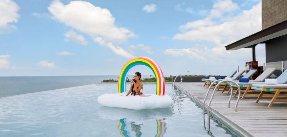 A person sits on a cloud-shaped inflatable with a rainbow arch in an infinity pool overlooking the ocean, with lounge chairs on the deck to the right under a partly cloudy sky.
