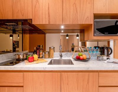 A clean, modern kitchen counter with a stainless steel sink, a black induction stovetop with a pot, various food items including bottles of oil and vinegar, jars of pasta, and a basket of fruit, all set against a backdrop of wooden cabinetry and a reflective panel.