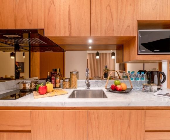 A clean, modern kitchen counter with a stainless steel sink, a black induction stovetop with a pot, various food items including bottles of oil and vinegar, jars of pasta, and a basket of fruit, all set against a backdrop of wooden cabinetry and a reflective panel.