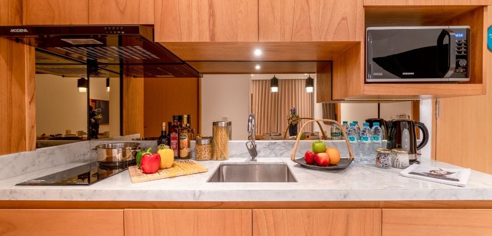 A clean, modern kitchen counter with a stainless steel sink, a black induction stovetop with a pot, various food items including bottles of oil and vinegar, jars of pasta, and a basket of fruit, all set against a backdrop of wooden cabinetry and a reflective panel.