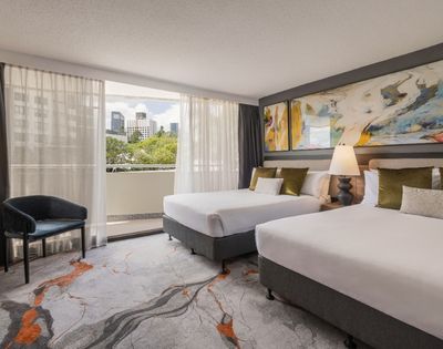 Hotel room with two beds, a chair, a large abstract painting on the wall, and a view of a city skyline through a balcony door.