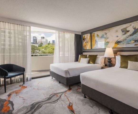 Hotel room with two beds, a chair, a large abstract painting on the wall, and a view of a city skyline through a balcony door.