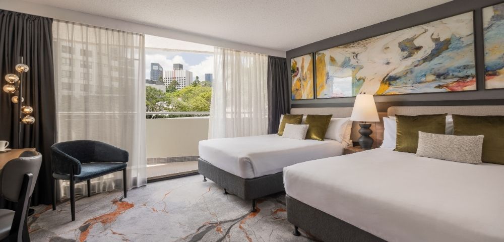 Hotel room with two beds, a chair, a large abstract painting on the wall, and a view of a city skyline through a balcony door.