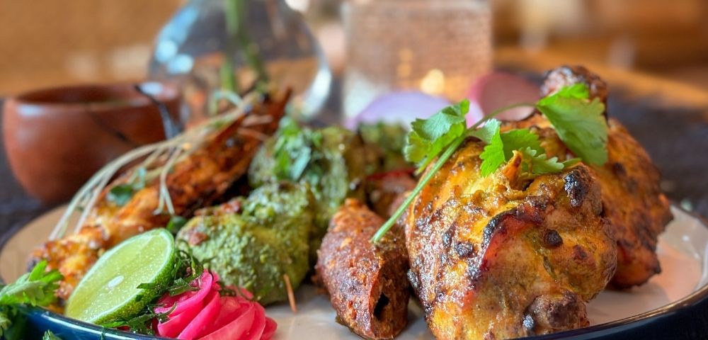A platter of Indian grilled food, including tandoori chicken, kebabs, and prawns, garnished with lime, pickled onions, and cilantro.