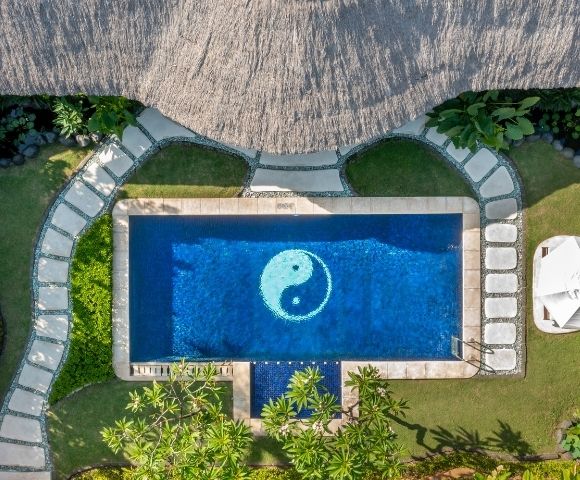 Aerial view of a rectangular swimming pool with a white yin and yang symbol at the bottom, surrounded by lush green grass, a thatched roof structure, and a stone pathway.
