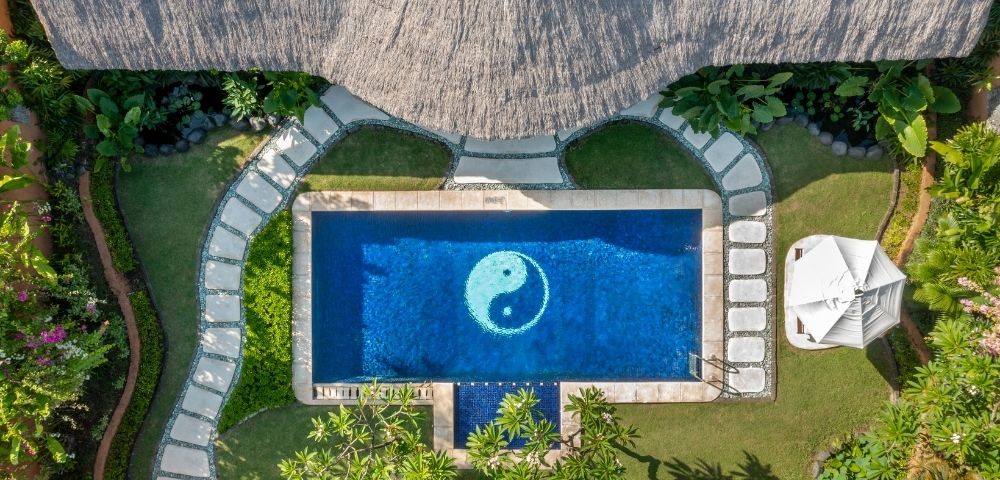 Aerial view of a rectangular swimming pool with a white yin and yang symbol at the bottom, surrounded by lush green grass, a thatched roof structure, and a stone pathway.
