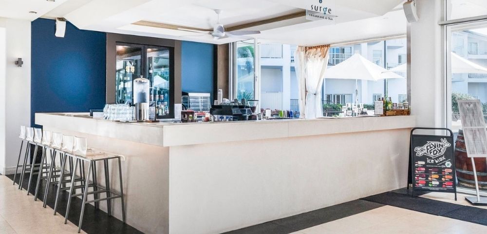 A bright, modern bar with a white counter, bar stools, and a dark blue accent wall, featuring a 