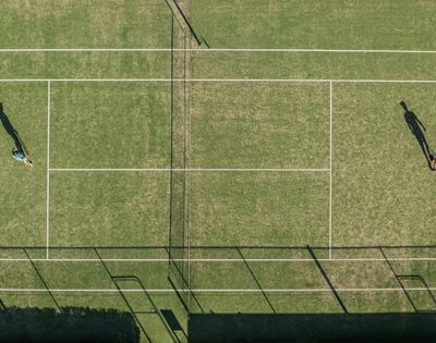 Aerial view of a grass tennis court with two people playing a match, casting long shadows.