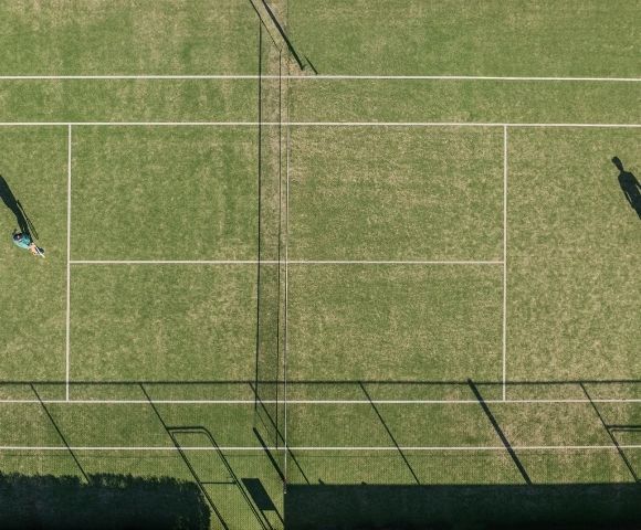 Aerial view of a grass tennis court with two people playing a match, casting long shadows.