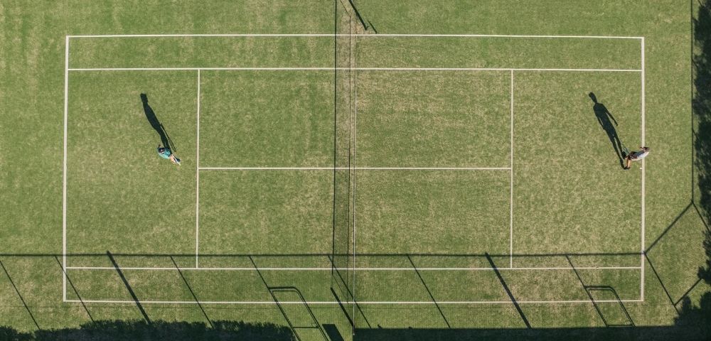 Aerial view of a grass tennis court with two people playing a match, casting long shadows.