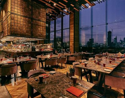Modern restaurant interior at dusk with marble tables, plush chairs, large windows showing city skyline, and a central bar area.