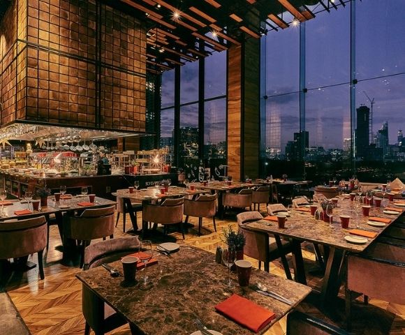 Modern restaurant interior at dusk with marble tables, plush chairs, large windows showing city skyline, and a central bar area.