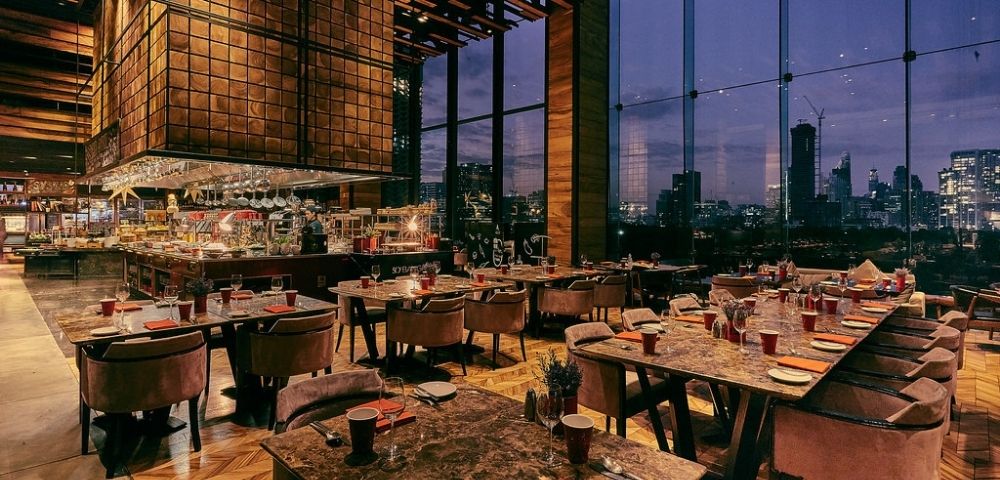 Modern restaurant interior at dusk with marble tables, plush chairs, large windows showing city skyline, and a central bar area.