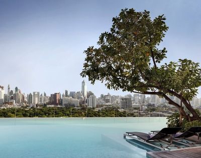 Infinity pool with lounge chairs and tree, overlooking a city skyline with tall buildings under a clear sky.