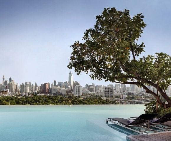 Infinity pool with lounge chairs and tree, overlooking a city skyline with tall buildings under a clear sky.