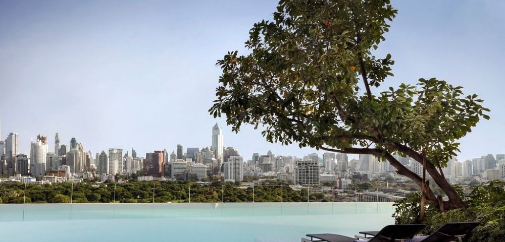 Infinity pool with lounge chairs and tree, overlooking a city skyline with tall buildings under a clear sky.