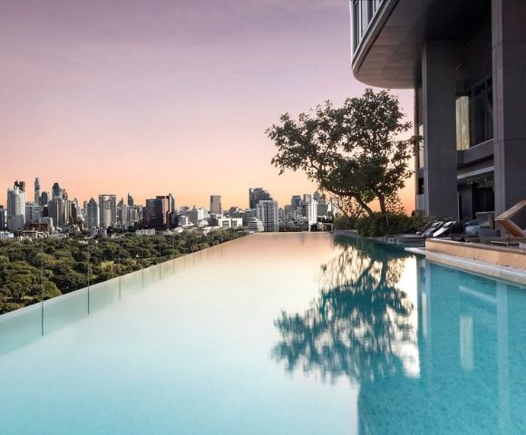 Infinity pool on a rooftop overlooking a city skyline at sunset with trees and lounge chairs nearby.
