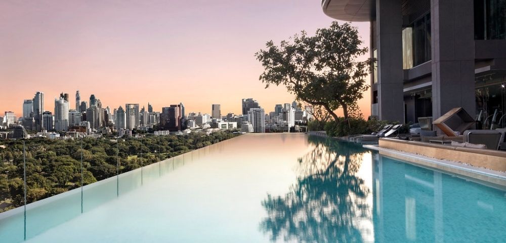 Infinity pool on a rooftop overlooking a city skyline at sunset with trees and lounge chairs nearby.