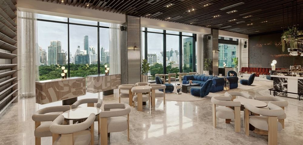 Spacious modern lounge with floor-to-ceiling windows, beige chairs, blue sofas, marble floors, and city skyline views in the background.
