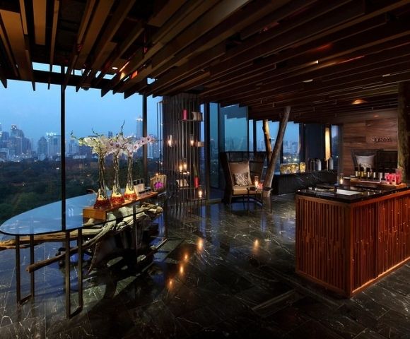 A modern interior of a spa or wellness center featuring sleek wooden decor, large windows showcasing a city view, and a reception area with candles and plants.